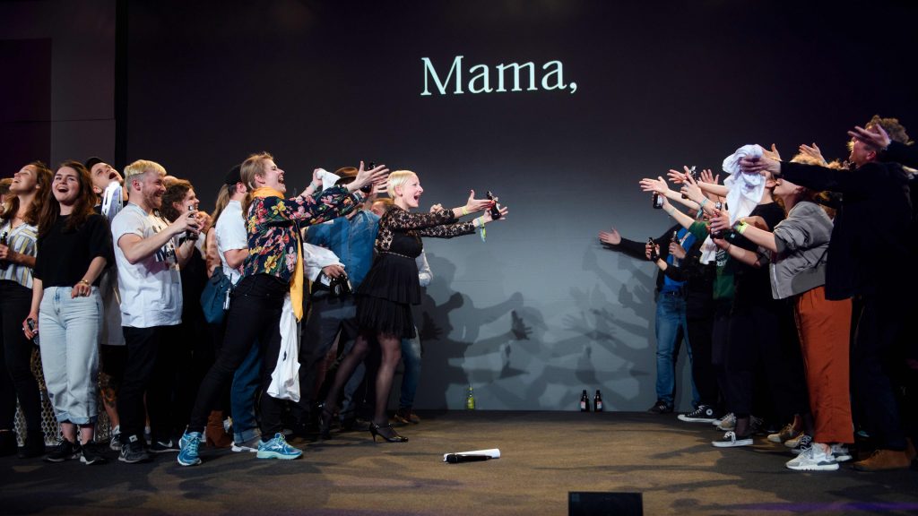 Menschen singend und posierend auf einer Bühne, auf deren Hintergrund „Mama“ steht